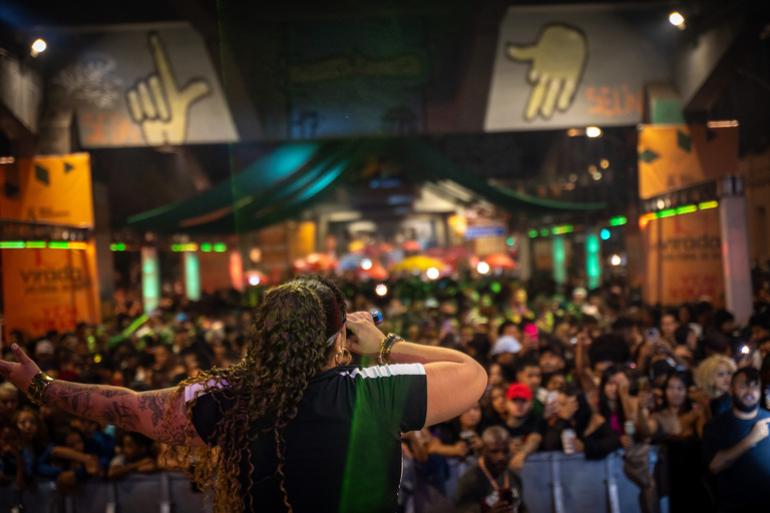 Virada Cultural de BH inicia avaliação das mais de 900 propostas inscritas Uma mulher de costas para a câmera, segurando um microfone, cantando e com um dos braços estendidos para a sua lateral. Ela está de frente para uma multidão em um local de evento, colorido, bem iluminado e com panos e pinturas no teto e nas laterais.