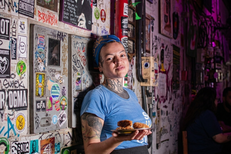 Viradão Gastronômico amplia experiência da Virada Cultural com circuito de bares e restaurantes no Centro Fotografia em plano médio de uma mulher jovem, parda, com cabelos cobertos por um lenço azul ornamentado e diversas tatuagens no pescoço e braços. Ela veste uma camiseta azul e segura um pequeno prato com bolinhos fritos empanados e dourados.