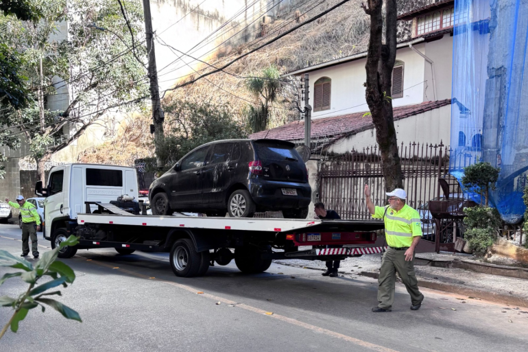 Mais de 1,7 mil veículos abandonados foram removidos das vias da cidade após intervenção da PBH Um caminhão-guincho branco está parado em uma rua íngreme e gramada para rebocar um carro preto danificado em uma área residencial arborizada. Dois agentes da BHTrans coordenam a operação, com um deles sinalizando na frente do veículo e o outro posicionado na parte traseira.