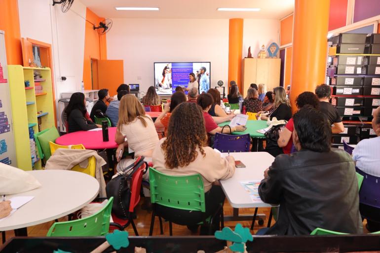 PBH e Uemg oferecem pós-graduação em Linguagem, Alfabetização e Letramentos para professores da rede municipal A foto mostra uma sala ampla e colorida, com várias mesas redondas e cadeiras de diferentes cores. Cerca de 20 pessoas estão reunidas, aparentemente participando de uma palestra ou treinamento, voltadas para uma televisão na parte da frente da sala. A tela exibe uma apresentação com imagens e textos. O ambiente possui paredes e colunas em tons de laranja, armários, estantes com materiais organizados e ventiladores nas paredes. A cena transmite um clima de reunião, aprendizado e interação.