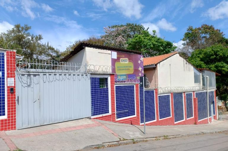 Fachada da Escola Municipal Lídia Angélica. O imóvel é térreo, com paredes revestidas por pastilhas nas cores azul, vermelho e branco e portões metálicos. A entrada e os muros são protegidos por concertina. Na fachada há uma placa de identificação e a logomarca da Prefeitura. O prédio possui telhados de diferentes alturas, janelas com grades e uma área de acesso com rampa de concreto. Em frente à escola, há uma calçada e a rua. Ao fundo, tem árvores de grande porte sob. céu azul com nuvens