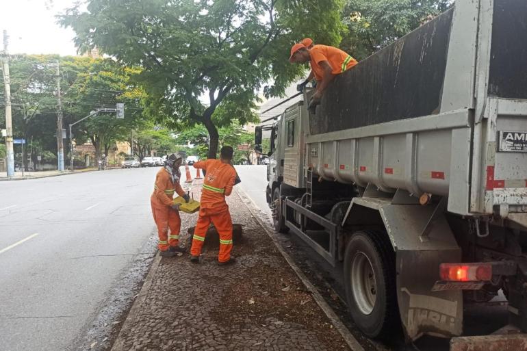A foto mostra três trabalhadores da limpeza urbana, vestidos com uniformes laranjas e faixas refletivas, realizando o recolhimento de resíduos na calçada. Dois profissionais estão no chão, manuseando os materiais, enquanto o terceiro está sobre a carroceria de um caminhão de coleta. O veículo ocupa boa parte do lado direito da imagem. A cena ocorre em uma via urbana arborizada, com calçada de pedras portuguesas, árvores e veículos ao fundo.