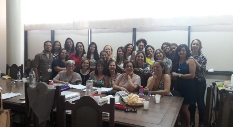 Projeto Appia Cerca de 20 pessoas posam para a foto, seis dela sentadas em cadeiras atrás de uma mesa de madeira, outras em pé.
