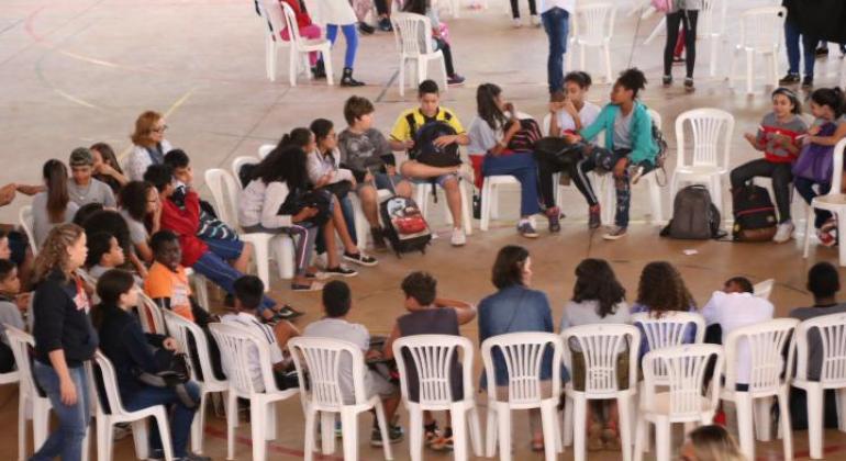 Grupo de adolescentes sentados em cadeiras brancas de plástico, em círculo, fazem uma roda de conversa na escola.