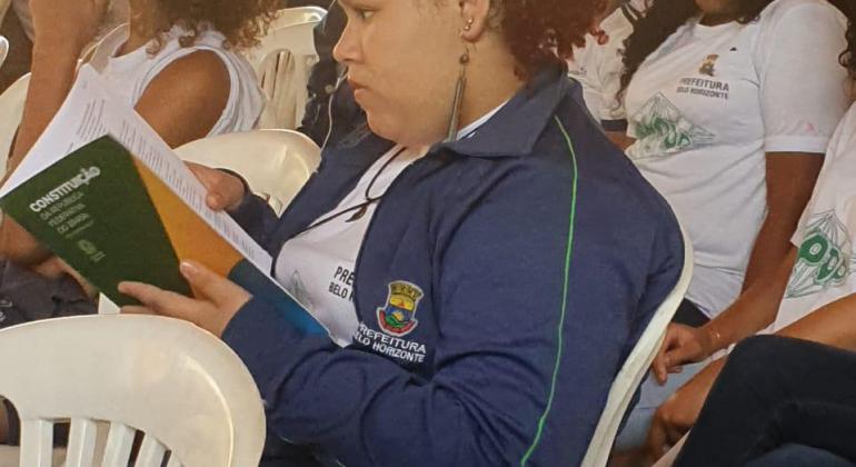 Aluna adolescente usa um uniforme da PBH, com camiseta branca e agasalho azul marinho, e lê um livro com capa verde e amarela.