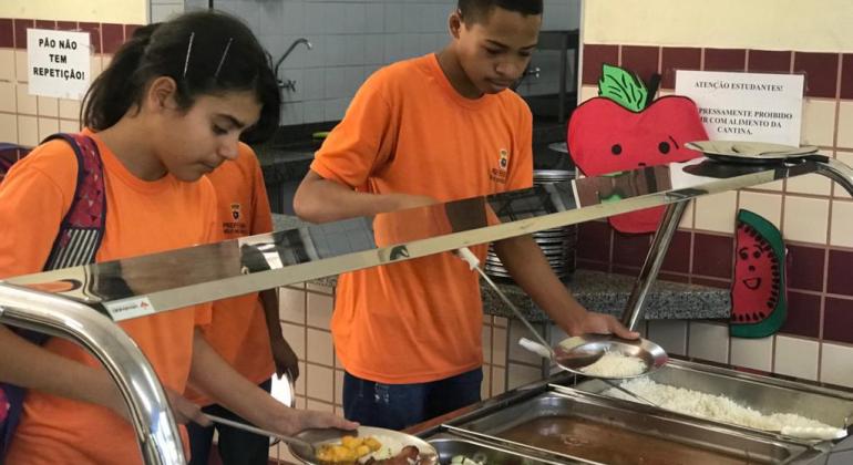 Dois alunos adolescentes , um menino e uma menina, usando camisetas cor de laranja estão em frente à um bandejão de uma cantina, servindo seus pratos.