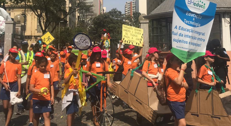 Alunos estão na rua vestidos de bermudas azul-marinho e camisetas cor de laranja, seguram cartazes, brinquedos e um estandarte, participando de um cortejo da educação.