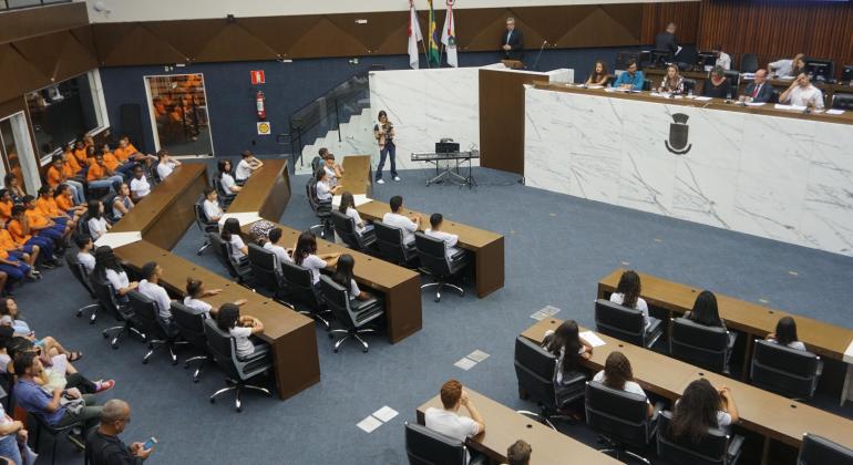 Câmara Mirim Alunos adolescentes sentam-se nas bancadas da Câmara dos Vereadores ouvindo vereadores que estão no plenário, à frente deles.