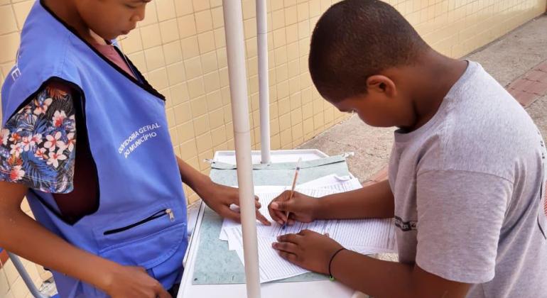 Um aluno vestido com um coleto azul claro, onde se lê: Ouvidoria Geral do Município, atrás de uma bancada indica o local de assinatura à uma outra criança, do lado da frente da bancada.