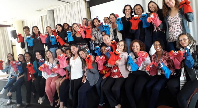 Cerca de 30 professoras posam para foto segurando um fantoche vermelho feito de feltro, representando o Projeto Vila Sésamo.