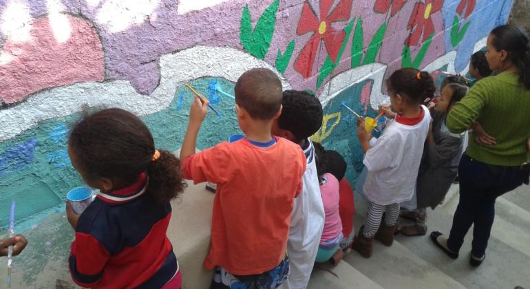 Cerca de 8 crianças e uma professora em uma escadaria. As crianças estão segurando pincéis e pintam um muro onde pode-se ver algumas flores vermelhas desenhadas