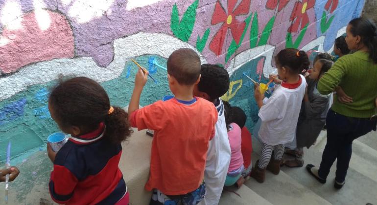 Projeto Rede Parceira em Movimento Cerca de 8 crianças e uma professora em uma escadaria. As crianças estão segurando pincéis e pintam um muro onde pode-se ver algumas flores vermelhas desenhadas