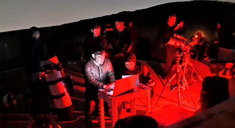 Cerca de oito pessoas estão em um ambiente escuro, iluminados por uma intensa luz vermelha e mexem em equipamentos eletrônicos.