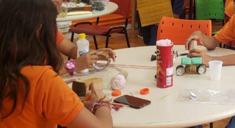Projeto Robótica e Programação Três crianças sentada em volta de uma mesa branca redonda, vestindo camisetas alaranjadas fazem trabalhos manuais usando materiais recicláveis como papelão, copo de plástico e outros