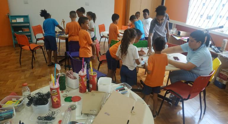 Doze crianças estão em uma sala com dois monitores. Eles estão de pé ao redor de mesas redondas brancas e cadeira alaranjadas. Eles estão fazendo trabalhos manuais com materiais como papelão, canudos plásticos, cola, canetas coloridas, garrafas plásticas.