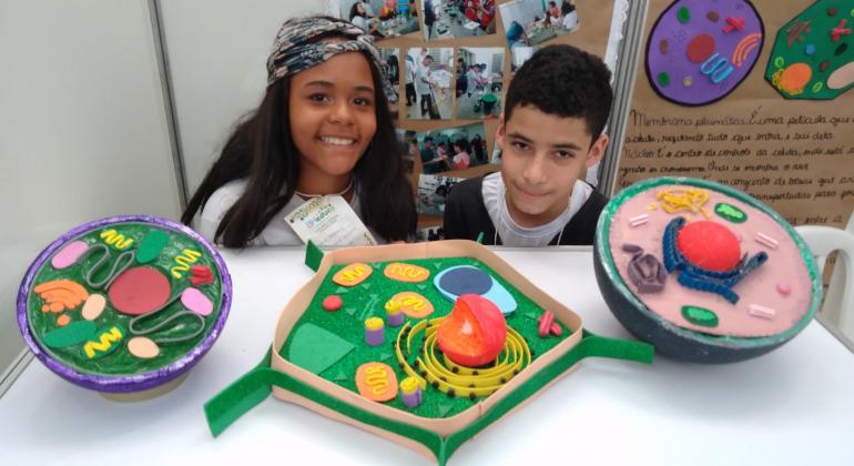 Uma menina e um menino sorriem para a foto, onde também se pode ver trabalhos feitos para a mostra científica, dois redondos e um em formato de pentágono.