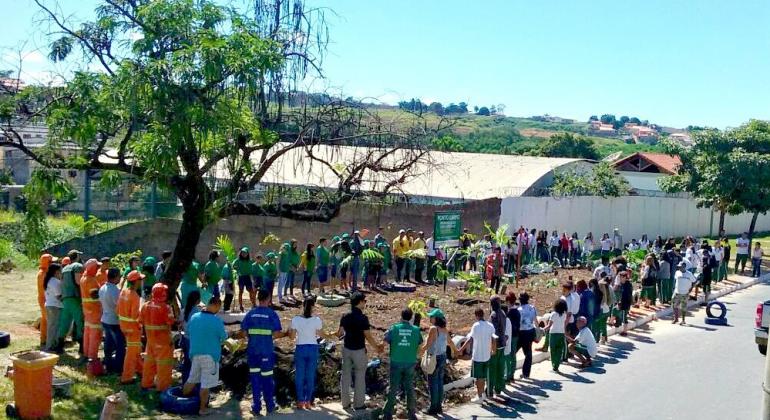 Várias pessoas de mãos dadas ao redor de um espaço arborizado com uma placa ao fundo