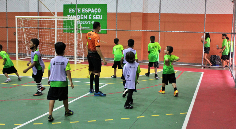 Futsal praticado crianças dentro de uma quadra poliesportiva. Presença de árbitro e de jogadores. 