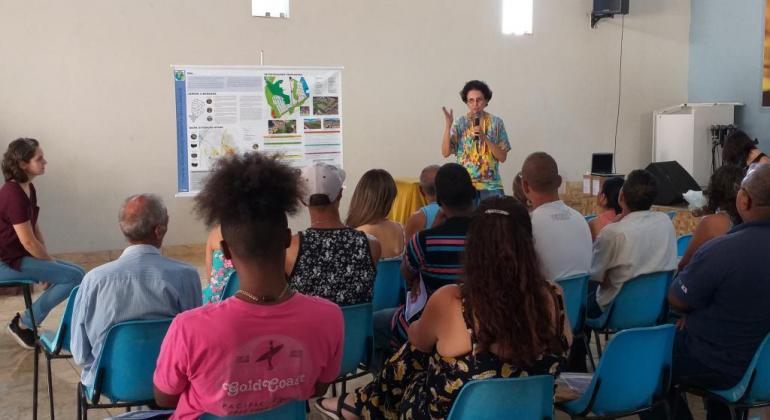 Em uma sala, pessoas assistem à apresentação do tema feita por uma mulher.