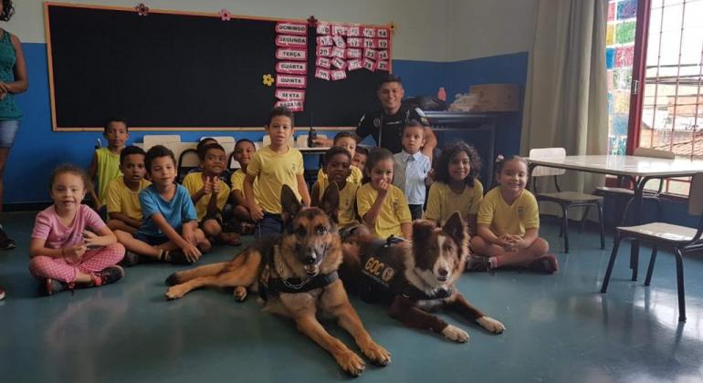 Cães da Guarda Municipal levam alegria às escolas da capital Em uma sala de aula, alunos e dois cães da Guarda Civil posam para a foto.
