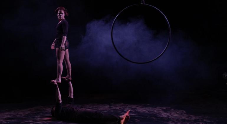 Teatro Francisco Nunes recebe espetáculo circense com acrobatas Em um palco, um artista deitado sustenta com as mãos uma artista. Ela está de pé, olhando para um aro suspenso por uma corda