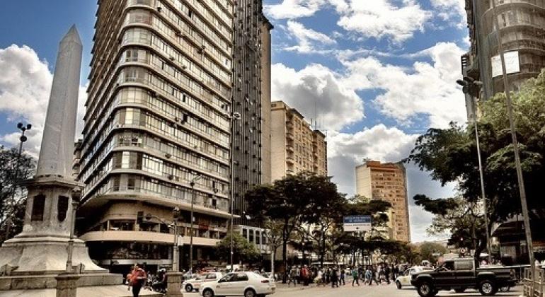 Praça Sete, no centro de Belo Horizonte, com um obelisco à esquerda e edifícios altos, rua e movimento pessoas e trânsito em um dia claro com nuvens.