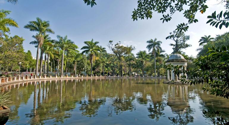 uma vista de uma lado da lagoa que fica no parque municipal Américo Renné Giannetti. Ela está rodeada por árvores e no meio dela tem um coreto. A foto foi tirada de dia, o céu está muio azul e sem nenhuma nuvem