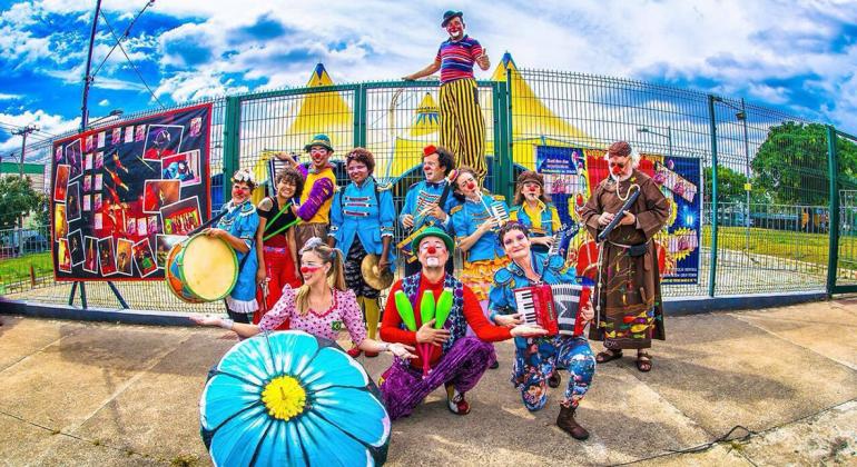 Doze membros de uma trupe de circo, com instrumentos musicais, malabares e outros acessórios, sorriem em frente à tenda de circo e à grade durante o dia. Foto: Divulgação