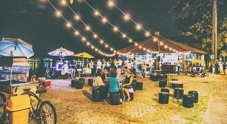 Cerca de vinte pessoas confraternizam ao lado do Centro de Referência Turística Álvaro Hardy - Veveco, durante a noite, na orla da Lagoa da Pampulha. Pessoas sentadas em mesas conversam, outras estão de pé, outras passeiam. Foto: Ricardo Laf