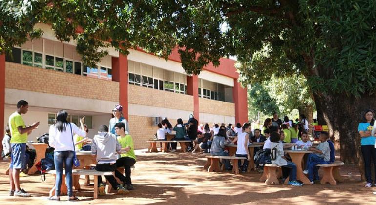 Mais de vinte alunos sentados em pátio de escola, com árvores e chão de terra e mesas e bancos em ambiente agradável durante o dia. Foto: Cláudio Lacerda