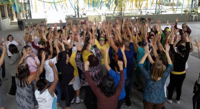 Centros Culturais oferecem oficinas de dança para idosos Mais de trinta idosos e adultos sorriem agrupados com as mãos para cina. Foto: Camila Mageste
