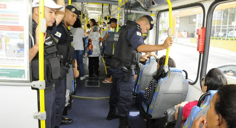 Três guardas municipais acompanham viagem de ônibus durante o dia. 