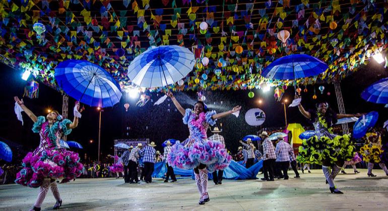 Três mulheres integrantes do grupo Feijão Queimado dançam com roupas típicas e sombrinhas, mais de seis homens dançam ao fundo e bandeirolas decoram o céu, penduradas por barbantes. 