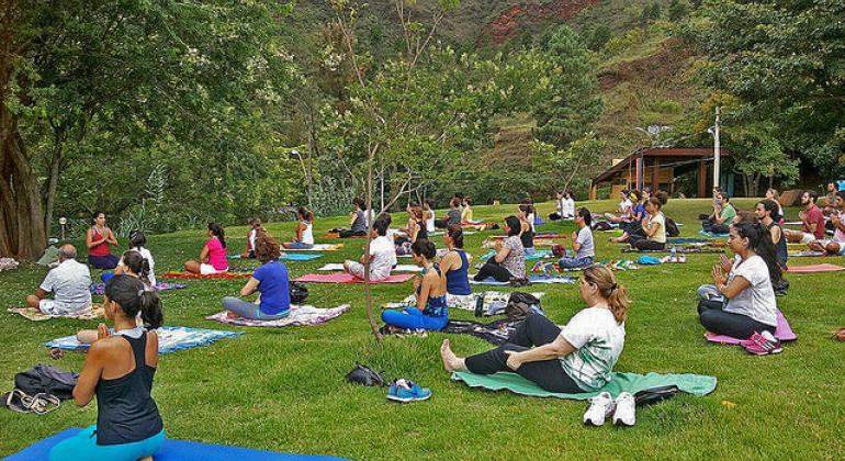 Aula de yoga no parque. Foto: Marta Amélia Cerca de 28 pessoas praticam yoga em um parque municipal, ajoelhadas em seus tapetes, durante o dia.