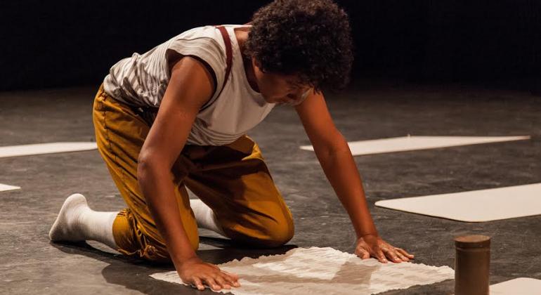 Criança ajoelhada com as mãos no chão olha para papel em branco; cena da peça infantil "O Pequeno". Foto: Filipe Hott