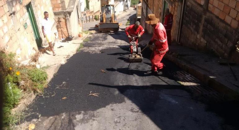 Beco com dois homens trabalhando com material e máquina tapa buracos.