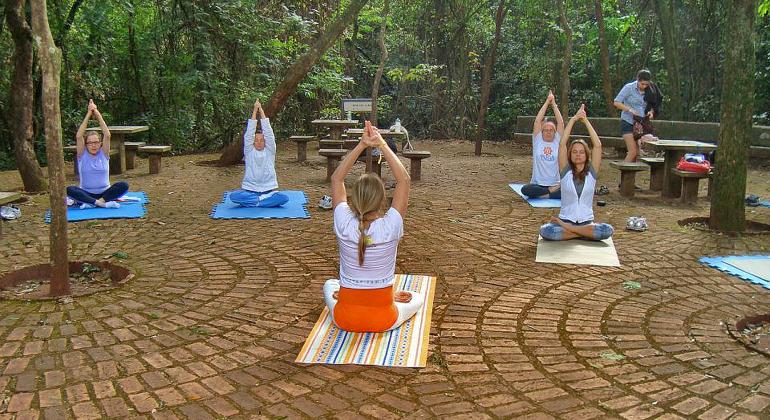 BH ao Ar Livre - Foto: Amyoga Mulheres sentadas em cima de toalhas com as mãos em cima da cabeça fazendo sinal de oração.