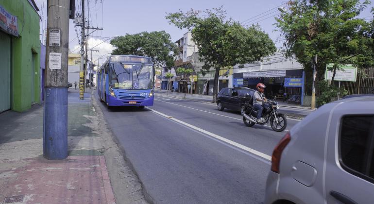 Na foto consta a traseira de um carro cinza em uma avenida, ao lado um motoqueiro em cima de uma moto e bem atrás um ônibus azul