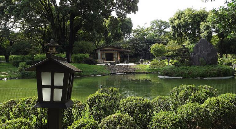 Jardim Japonês na Fundação Zoo-Botânica de BH, com arbustos redondos e uma lantarna à frente, um lago no centro e árvores e uma contrução atrás. 