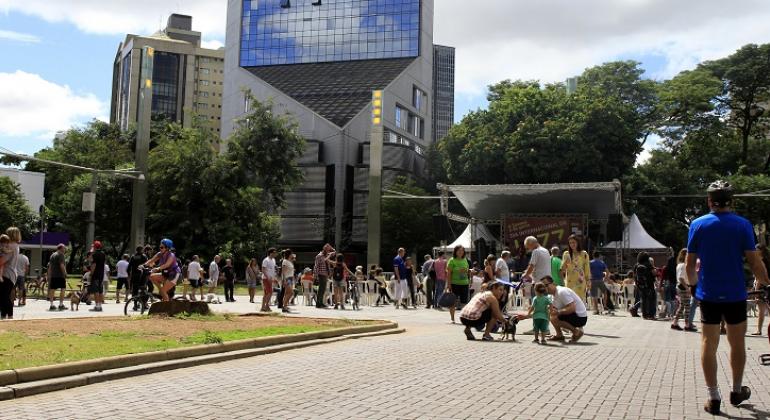 Programa “A Savassi é da Gente” volta com música e esporte - Foto: Amira Hissa Praça da Savassi movimentada no domingo. Cerca de cem pessoas na imagem. Alguns cidadãos praticam esportes, outros caminham e outros estão sentados em frente ao palco, montado na praça. Ao fundo, prédios que cercam a praça.
