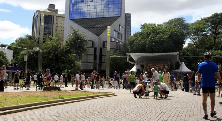 Praça da Savassi movimentada no domingo. Cerca de cem pessoas na imagem. Alguns cidadãos praticam esportes, outros caminham e outros estão sentados em frente ao palco, montado na praça. Ao fundo, prédios que cercam a praça. 