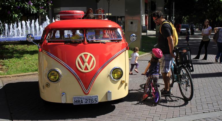 Homem e criança, cada um em uma bicicleta, observam, parados kombi amarela e laranja durante o dia. Ao fundo, a fonte sa praça da Savassi jorra água. 