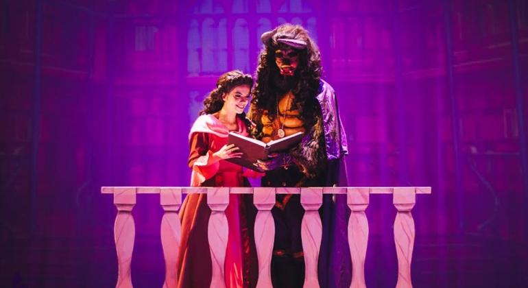 Bela, jovem com trajes de princesa, lê livro junto à Fera em uma sacada em cena teatral.