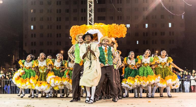 Mais de vinte homens e mulheres com trajes juninos em verde e amarelo fazem formação de quadrilha junina. 