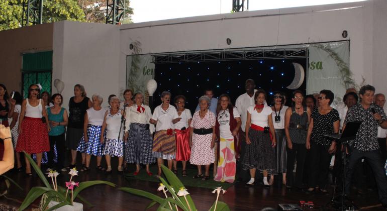 Cerca de vinte idosos caracterizados ao estilo da década de 1960, se apresentam no palco da homenagem ao Dia das Mães do Centro de Referência da Pessoa Idosa.