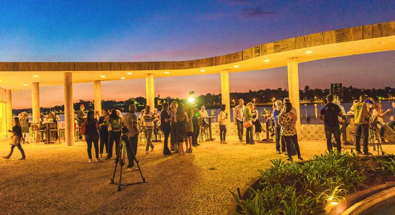 Cerca de 30 pessoas conversam entre si, confraternizam e olham para o ambiente na entrada da Casa do Baile à noite. À frete, observa-se um tripé com máquina fotográfica. 