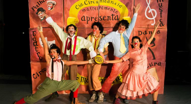 Cia. Circunstância apresenta peça “De Mala às Artes” dia 21 de maio no Teatro Francisco Nunes. Foto: Lina Mintz Quatro homens e uma mulher, com maquiagem de palhaço e roupas estilizadas, posam atrás de uma cartaz com os dizeres: Cia. Circunstância matreiramente apresenta