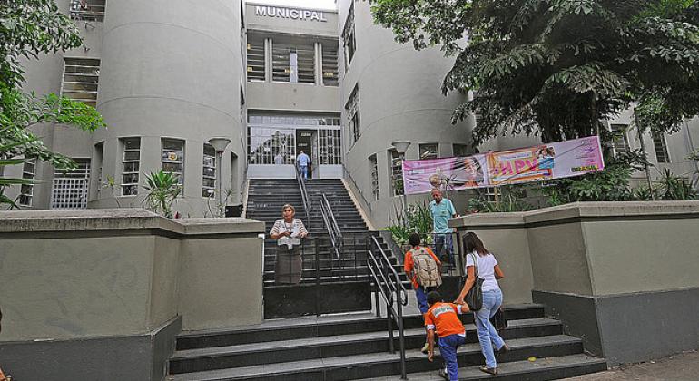 Fachada da Escola Municipal Marconi, que tem uma ampla escadaria, com cinco pessoas entrando e saindo durante o dia.