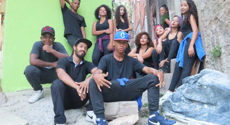 Grupo Identidade, formado por 10 jovens, homens e mulheres, todos de roupas pretas como detalhes como tênis, bonés e blusas, em azul, branco ou roxo. 