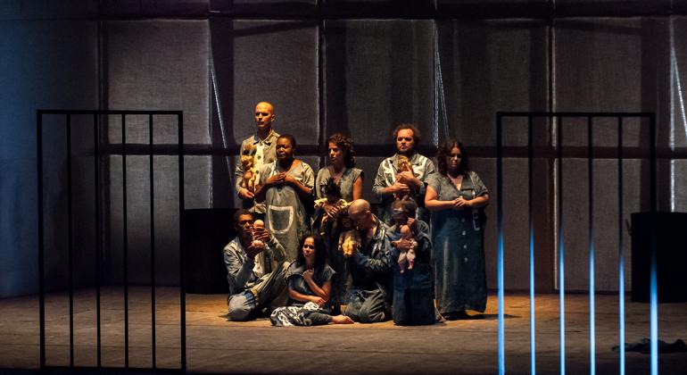Sete homens e mulheres com trajes cinza-azulados, alguns deles com bonecas, olham por trás de uma grade cênica, cortada ao meio, em um palco de teatro. 